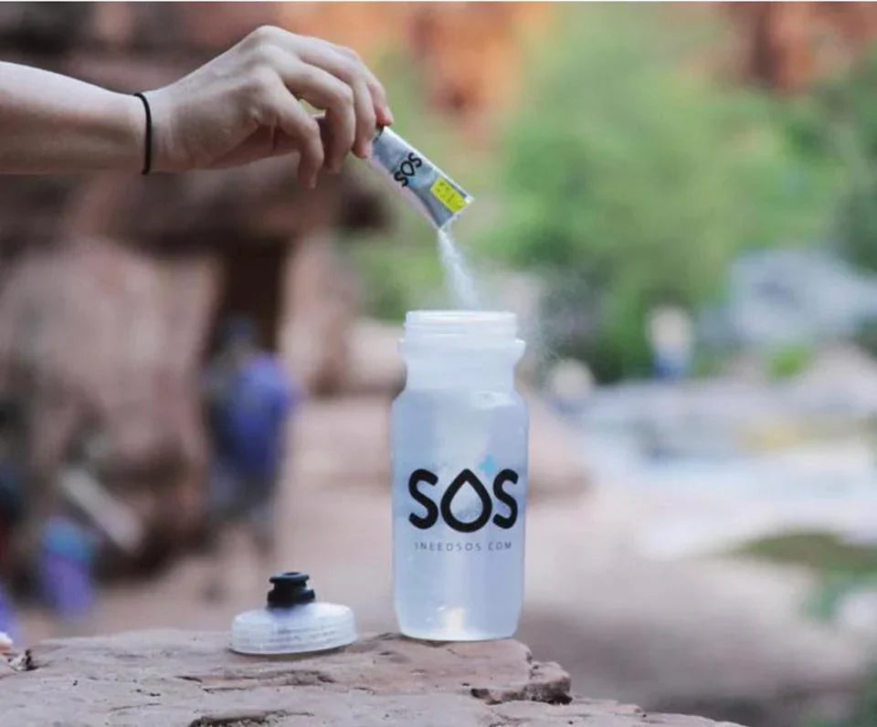 Person pouring SOS Hydration electrolyte powder into a water bottle outdoors with a rocky, nature background