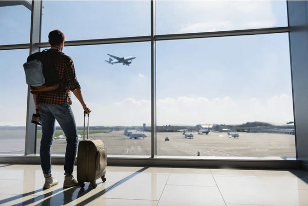 Traveler standing at airport terminal window with luggage, watching airplane take off on a clear day