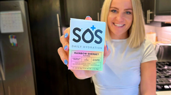 Smiling woman holding a box of SOS Daily Hydration Rainbow Sherbet electrolyte drink mix in a modern kitchen, promoting hydration and wellness.