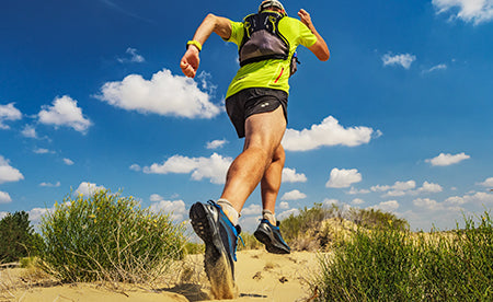 Athlete running on a rugged, sunlit trail with a hydration vest, captured mid-stride against a bright blue sky and scattered clouds.
