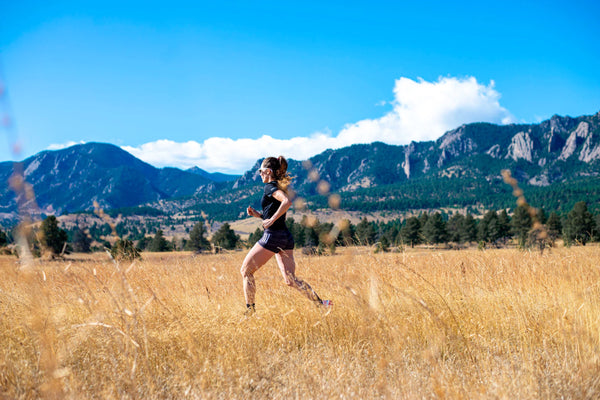 Female runner sprinting through a golden field with mountain views in the background on a clear, sunny day—symbolizing endurance, hydration, and outdoor fitness